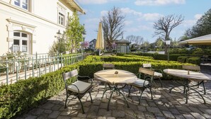 Un patio con mobiliario exterior, un edificio de arquitectura clásica y setos bien cuidados.
