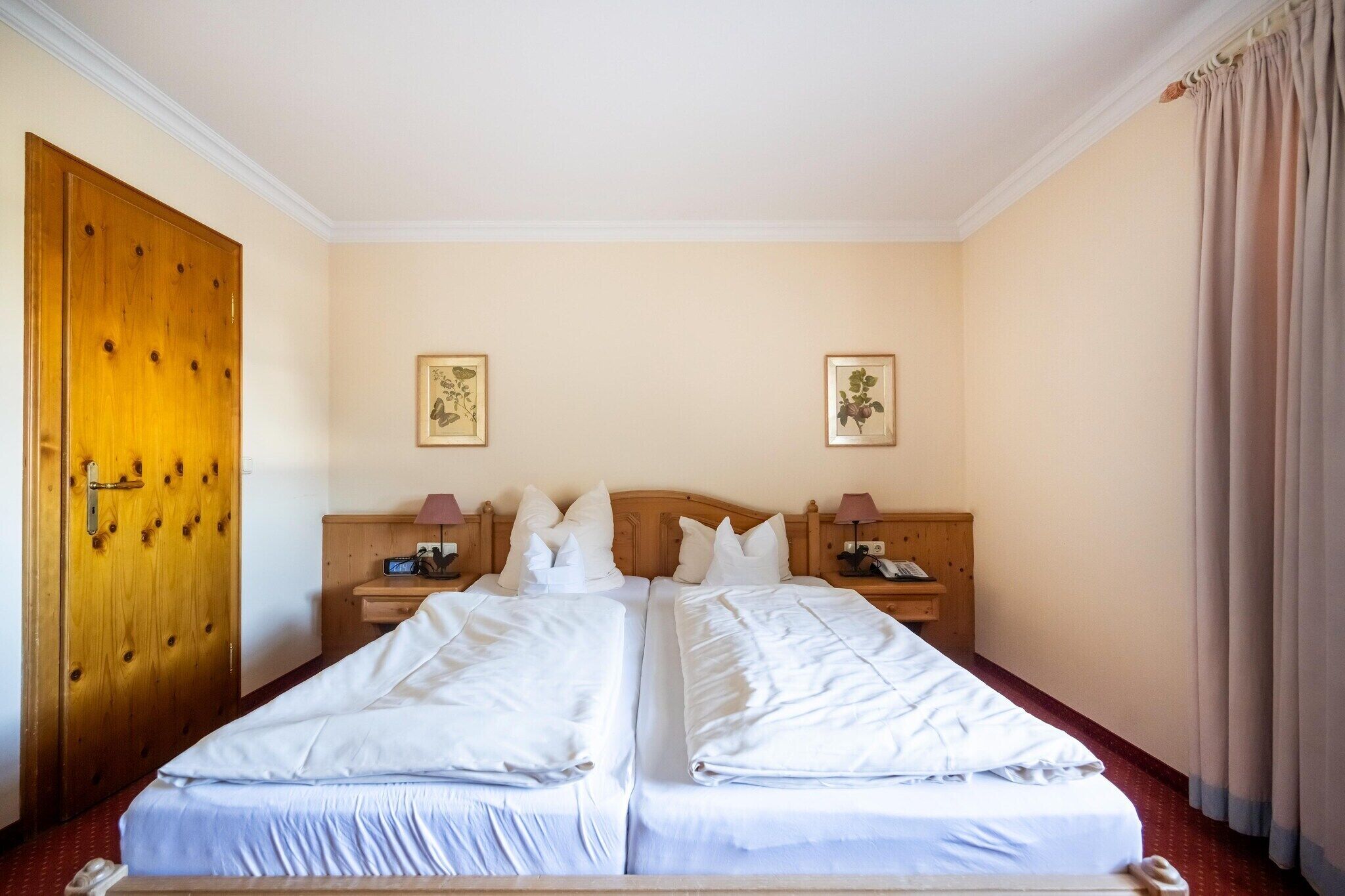 Une chambre d’hôtel avec deux lits, une tête de lit en bois, des tables de chevet avec des lampes et des œuvres d’art florales encadrées accrochées aux murs.