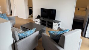 Un salon moderne avec des canapés gris, une table basse et un téléviseur à écran plat. On aperçoit une machine à laver à l’arrière-plan, ce qui indique la présence d’un espace buanderie.