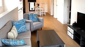 Un salon moderne avec un canapé gris, une table basse et un téléviseur à écran plat. On aperçoit à l’arrière-plan un coin repas avec une table et des chaises.