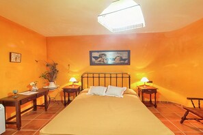 A bedroom with a bed, two bedside tables, a nightstand, a lamp, a potted plant, and a framed picture on the wall.