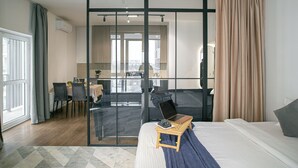 A modern hotel room with a large bed, a laptop on a bedside table, and a view through a sliding door to a dining area.