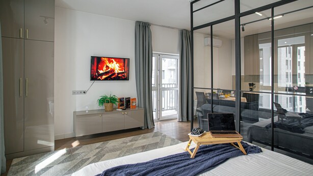A modern bedroom with a bed, a laptop on a small table, a TV displaying a fireplace, and a large window with curtains.