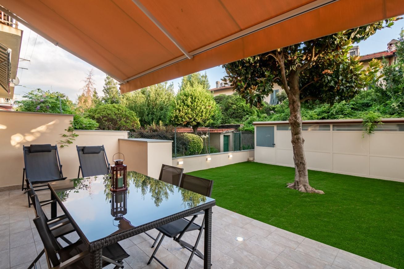 An outdoor patio with a glass table and chairs, surrounded by a well-maintained lawn and a tree.
