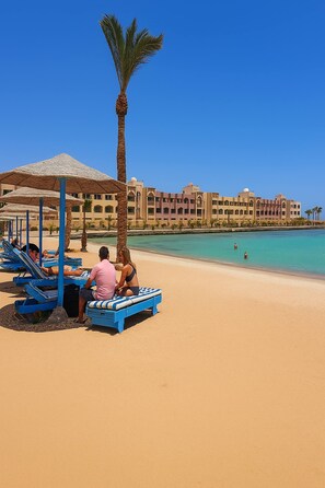Un hôtel situé en bord de mer, avec des chaises longues et des parasols, un palmier et un ciel bleu azur.