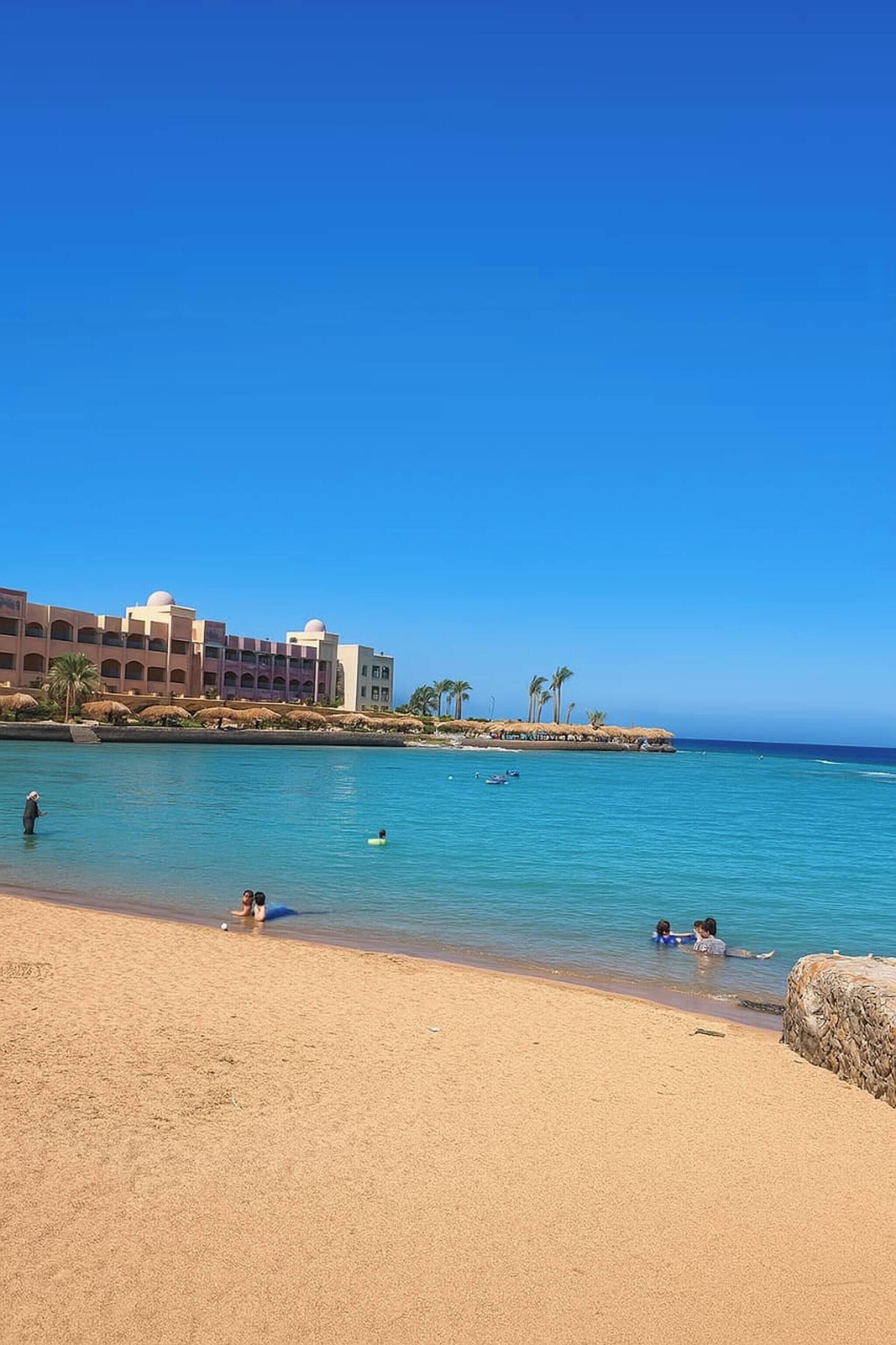 Ein Strand mit einem Hotel, klarem, blauem Wasser und einem sandigen Ufer.