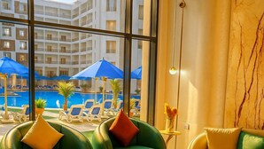 A modern hotel lobby with green leather sofas, a yellow sofa, and a glass coffee table. Large windows offer a view of a pool area with umbrellas and lounge chairs.