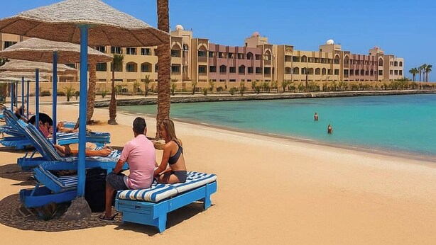 A beach with sun loungers and umbrellas, a palm tree, and a hotel in the background.