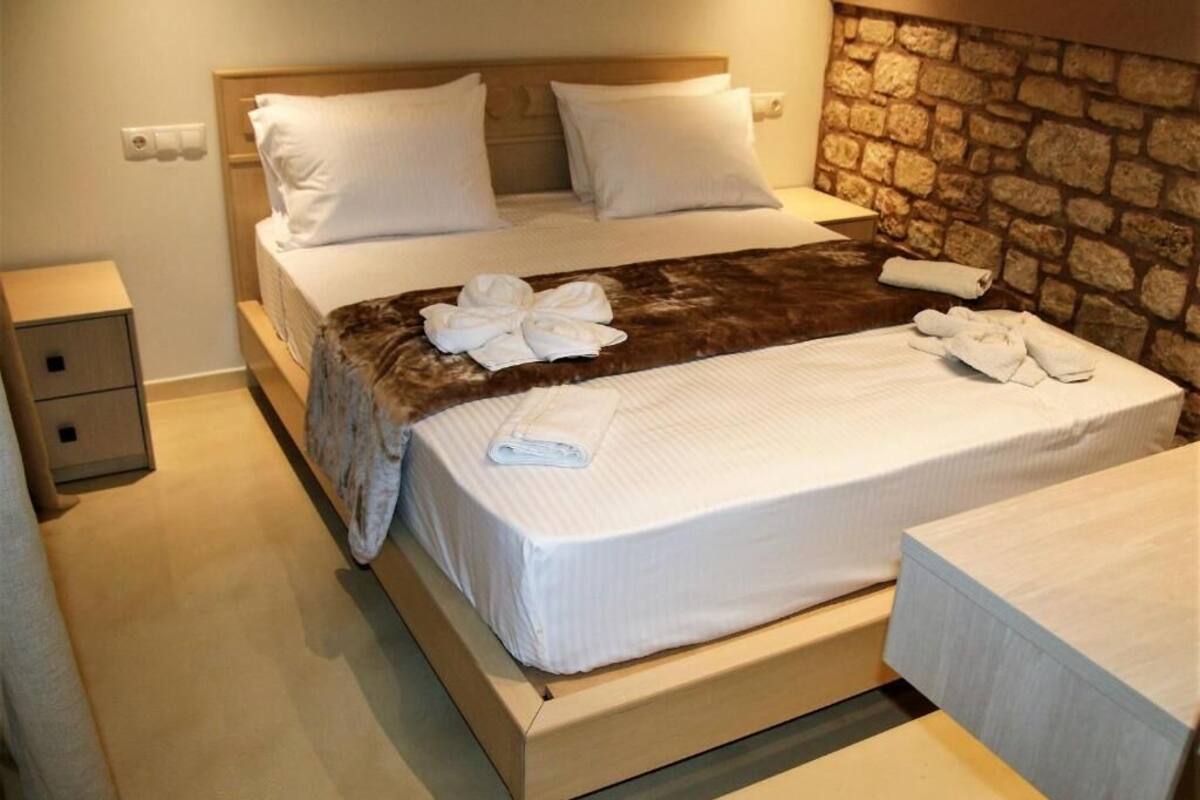 A neatly made bed with white linens and a brown throw blanket, a wooden headboard, a stone wall, and a small bedside table.