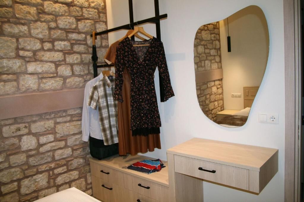 A room with a stone wall, a wooden dresser, a mirror, and a clothing rack with a dress and a plaid shirt.