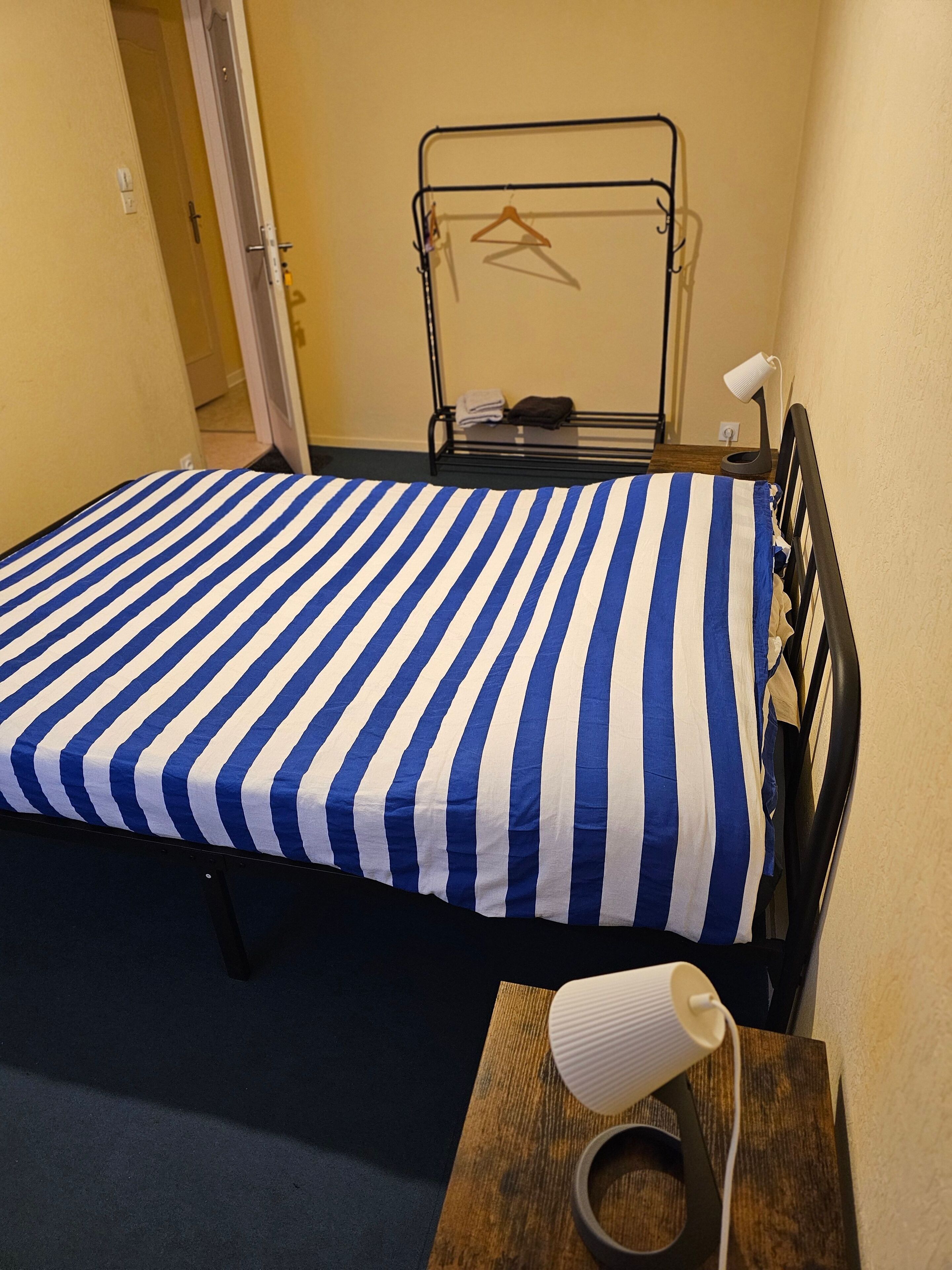 A small, neatly made bed with a blue and white striped bedspread in a room with a wooden nightstand, a lamp, and a coat rack with hangers.