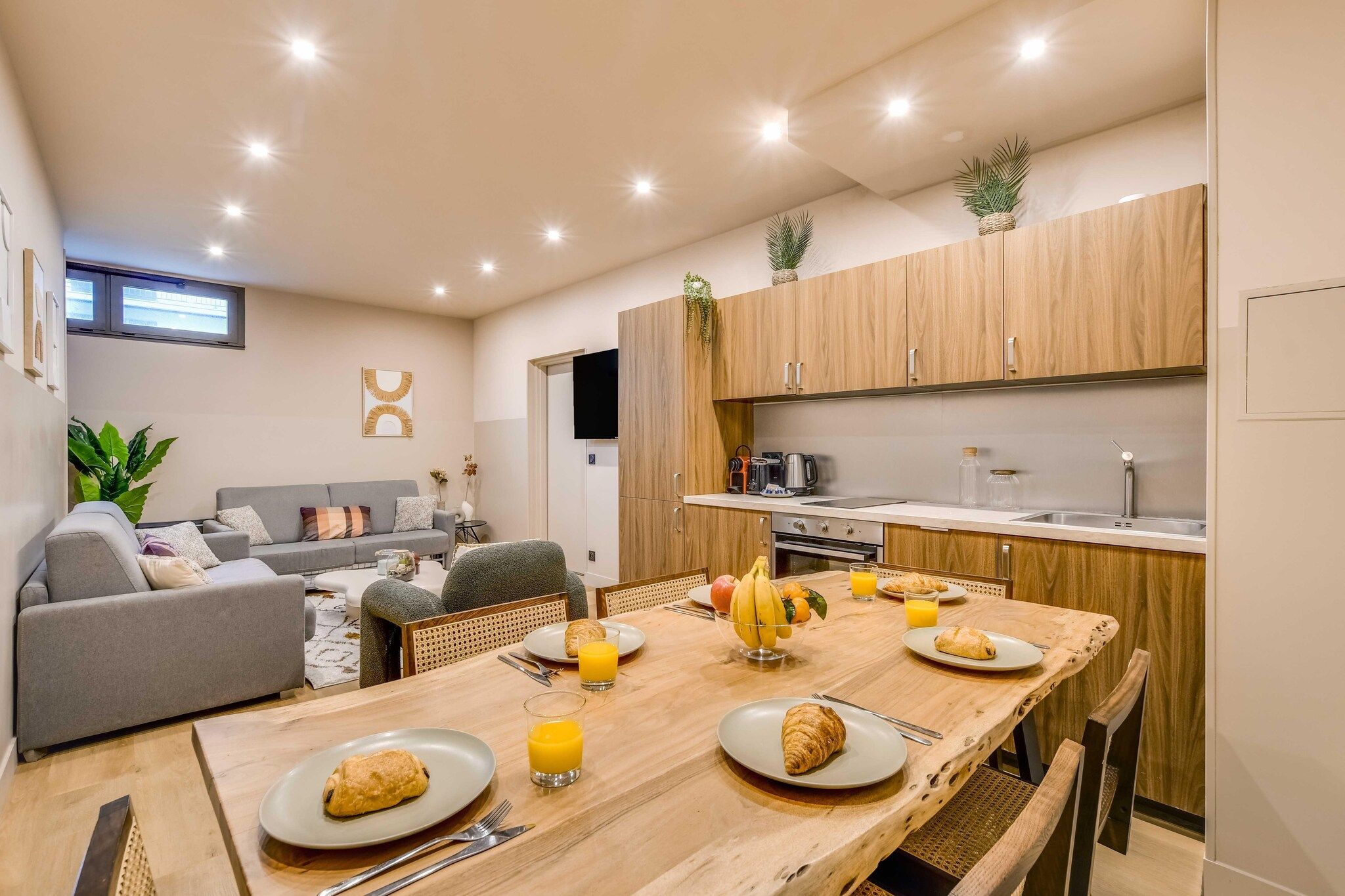 A modern kitchen with wooden cabinets, a dining table set with breakfast, and a living area with a sofa and plants.
