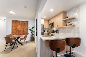 Una cocina moderna con armarios de madera, encimera de mármol y microondas integrado. Hay un comedor con mesa de vidrio y sillas marrones.