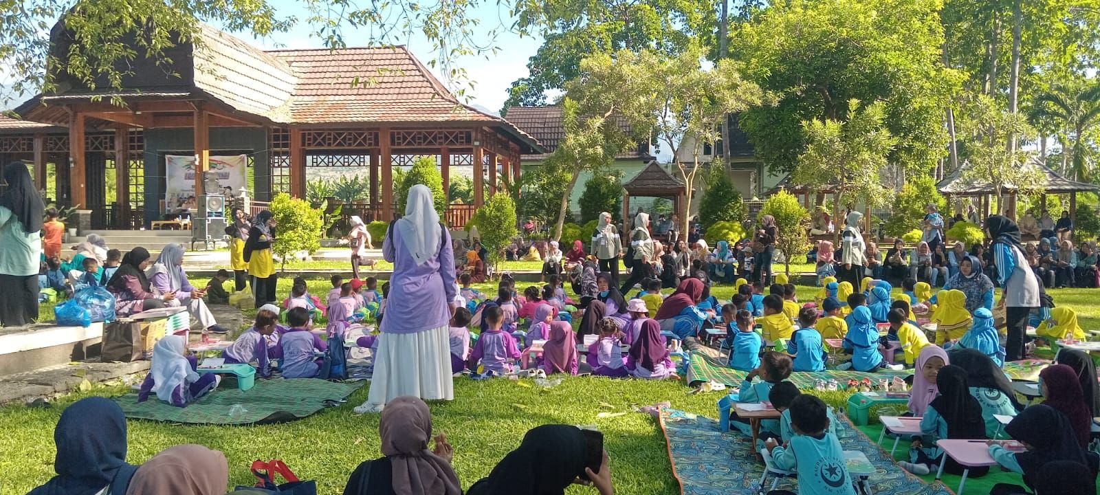 Une réunion en plein air où les participants sont assis sur l’herbe, certains sont debout, et on aperçoit une structure en arrière-plan.