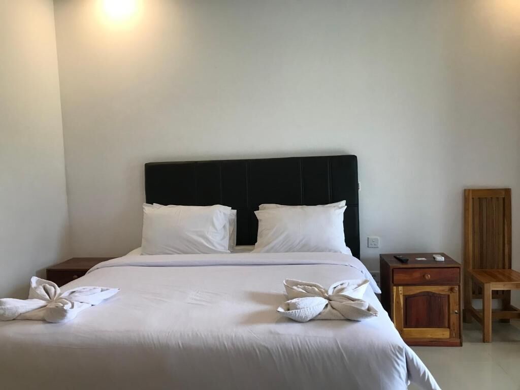 Un lit bien fait, avec du linge de lit blanc et des oreillers, une table de chevet en bois avec une télécommande, et une chaise en bois adossée au mur.