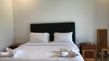 Un lit bien fait, avec du linge de lit blanc et des oreillers, une table de chevet en bois avec une télécommande, et une chaise en bois adossée au mur.