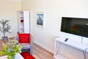 A modern living room with a grey sofa, red cushions, a white TV stand, and a potted plant. - Apartment Am Yachthafen auf Fehmarn (Fehmarn OT Burgtiefe-Südstrand)