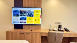 Ett hotellrum med en stor platt-TV som visar hotellets informationssida, ett skrivbord i trä med en telefon, ett glas vatten och en kopp.