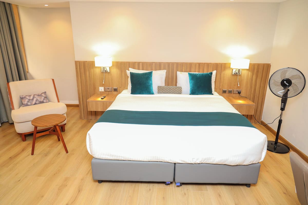 A hotel room with a large bed, two bedside tables with lamps, a chair, and a standing fan.