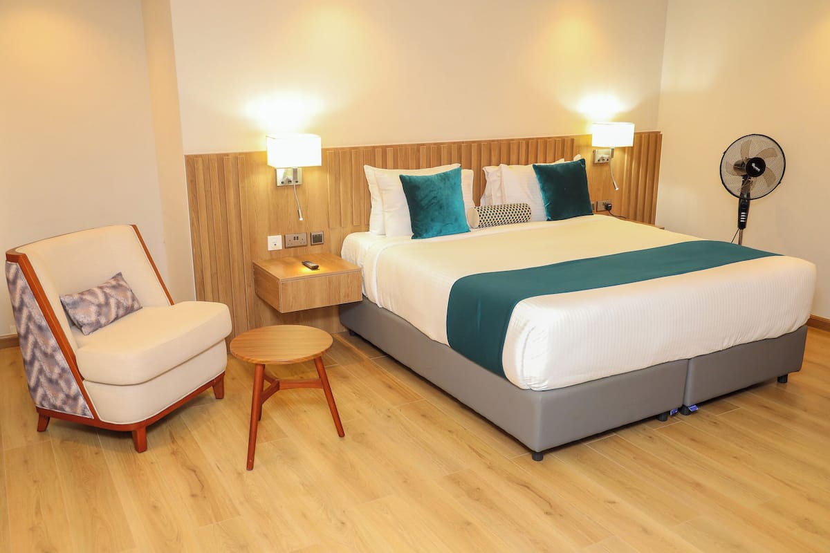 A hotel room with a bed, a chair, a small table, a nightstand, and a wall-mounted fan.