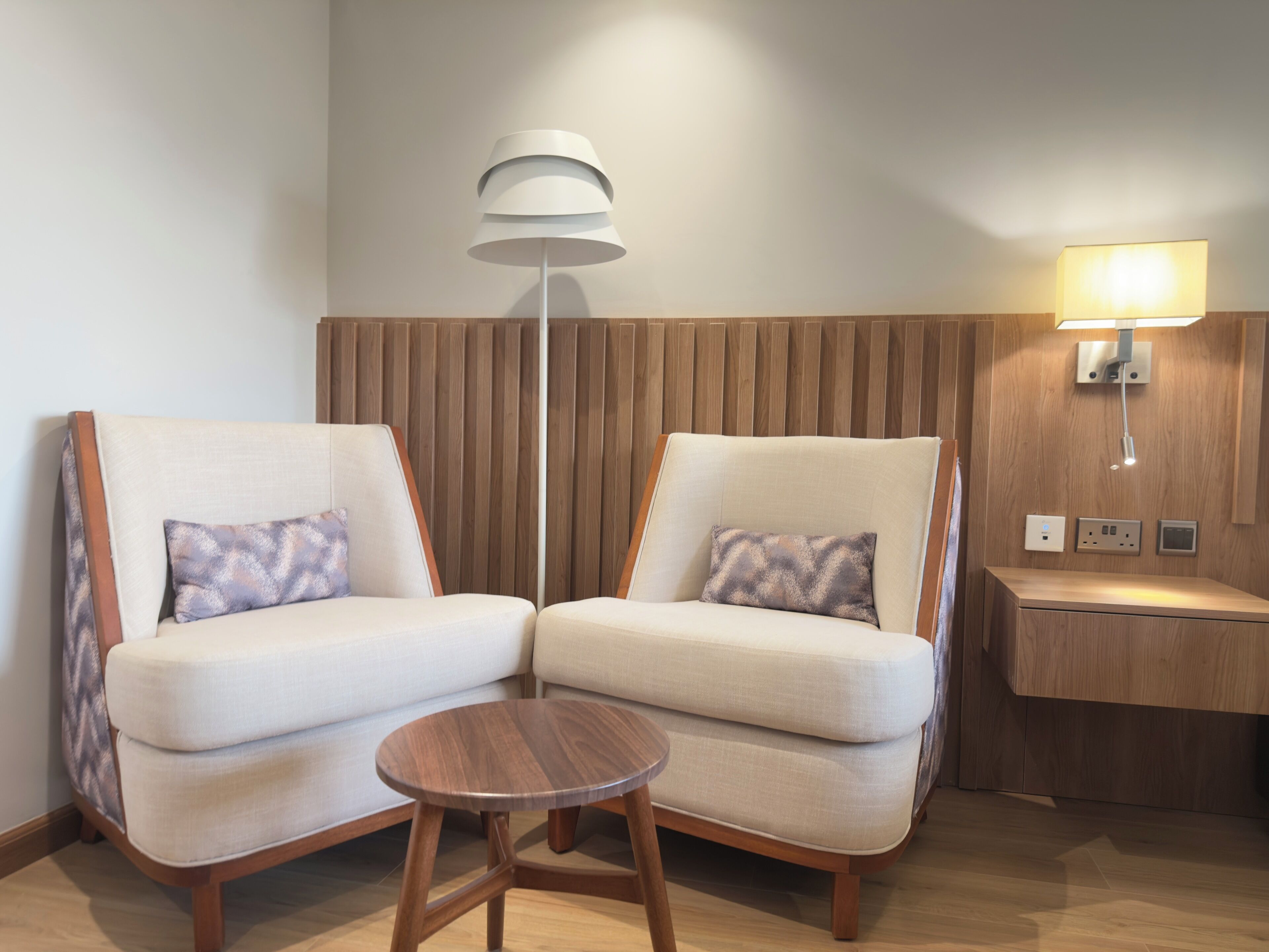 A modern hotel room with two sofas, a small wooden table, a floor lamp, and a bedside table with a lamp.