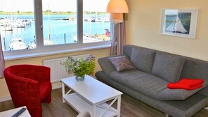 Un salón con un sofá gris, una silla roja, una mesa blanca y vistas a un puerto deportivo a través de amplios ventanales.