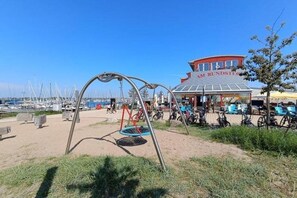 Ubicació a la platja amb un tobogan, un edifici amb la inscripció "AM RUNDSTEIG" i bicicletes aparcades a prop.