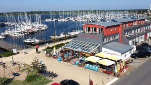 Un puerto deportivo con numerosas embarcaciones amarradas, un edificio rojo con escaparate de vidrio y una zona pavimentada con asientos y sombrillas.