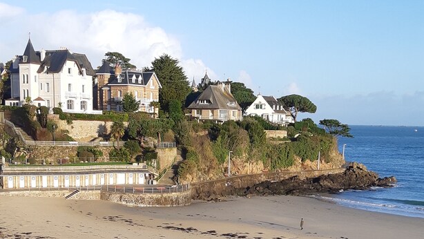 A coastal view with a balcony railing, a sandy beach, and houses with a stone wall and greenery.