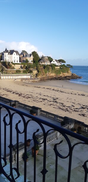 A coastal view with a balcony railing, a sandy beach, and houses with a stone wall and greenery.