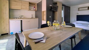 Une chambre d’hôtel compacte dotée d’une kitchenette, d’une table à manger dressée avec des assiettes, des couverts et un verre de vin, et d’un lit avec une tête de lit sombre.