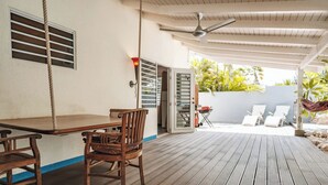 Um deck de madeira com mesa e cadeiras, um ventilador de teto e vista para uma área de piscina com espreguiçadeiras.