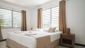 Quarto de hotel com uma cama grande, cortinas bege, uma mesinha de vime e vista pela persiana.