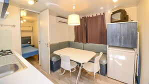 Een compacte kitchenette met een gasfornuis, een spoelbak en een kleine koelkast. Er is een eettafel met twee stoelen en een bank met kussens. Een televisie is aan de muur boven de bank bevestigd.