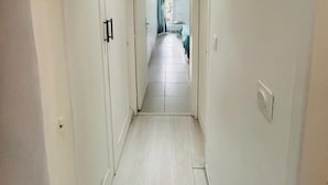 En smal hall med vita väggar och ett kaklat golv leder in till ett rum med en säng och ett fönster.