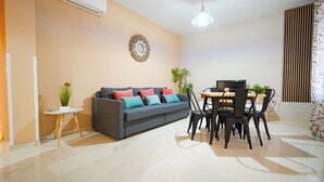 Een moderne woonkamer met een grijze bank, kleurrijke kussens, een klein bijzettafeltje met een plant, een eethoek met een houten tafel en zwarte stoelen, en een plafondlamp.