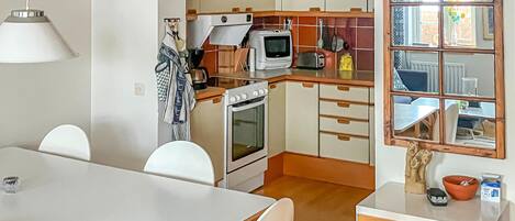 A compact kitchen with a stove, microwave, and wooden cabinets. There is a dining table with chairs and a window with a view of the outside.