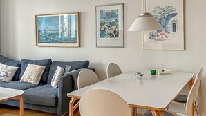 Uma sala de estar moderna com um sofá cinzento, uma mesa de jantar branca e cadeiras. Há obras de arte emolduradas na parede e uma luminária pendente sobre a mesa.