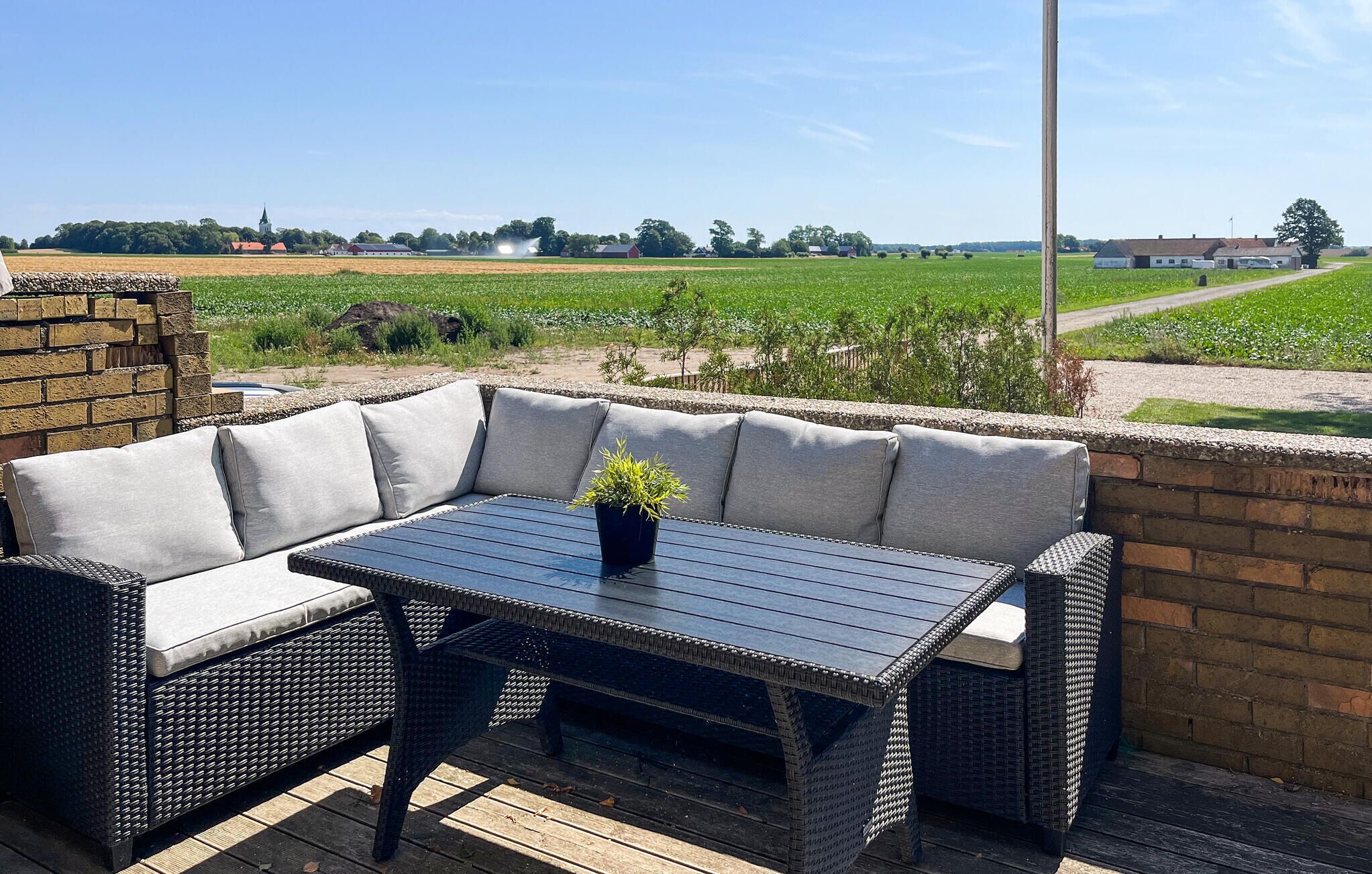 Un patio exterior con sofá y mesa de mimbre, una planta en maceta y vistas a un paisaje rural.
