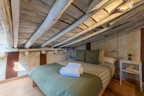 A bedroom with a stone wall, wooden ceiling beams, a bed with a green bedspread, and a bedside table with a lamp.