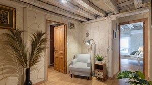 Ein Zimmer mit Holzboden, einem weißen Sessel, einer Vase mit Trockenpflanzen, einer Lampe und einer Holztür.