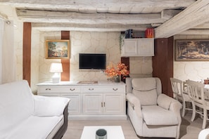 A living room with a white sofa, a beige armchair, a TV, a cabinet, and a dining table with chairs.