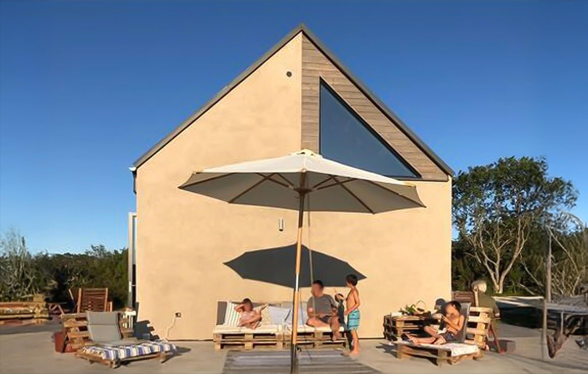 Ein großes, eingeschossiges Gebäude mit einem geneigten Dach, einem zentralen Innenhof mit Liegestühlen und einem Sonnenschirm sowie einem kleinen Poolbereich mit Liegestühlen und einem Tisch.