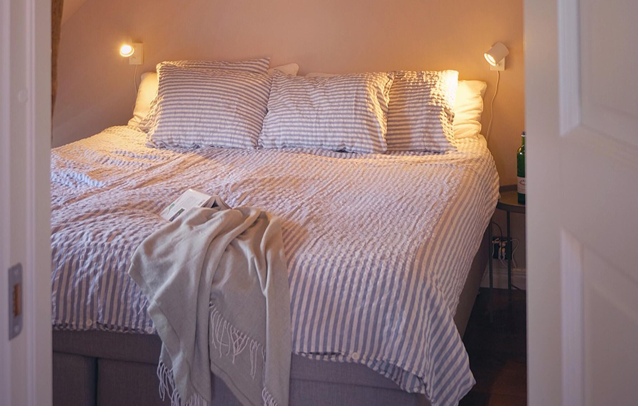 Un lit bien fait, avec une literie à rayures et des oreillers, une lampe de chevet et une bouteille de vin posée sur une petite table à côté du lit.