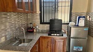 Eine Küche mit einer Granitabdeckung, einer Edelstahlspüle, Holzschränken, einem Gasherd, einem Kühlschrank und einem Fenster mit einem Metallgitter.