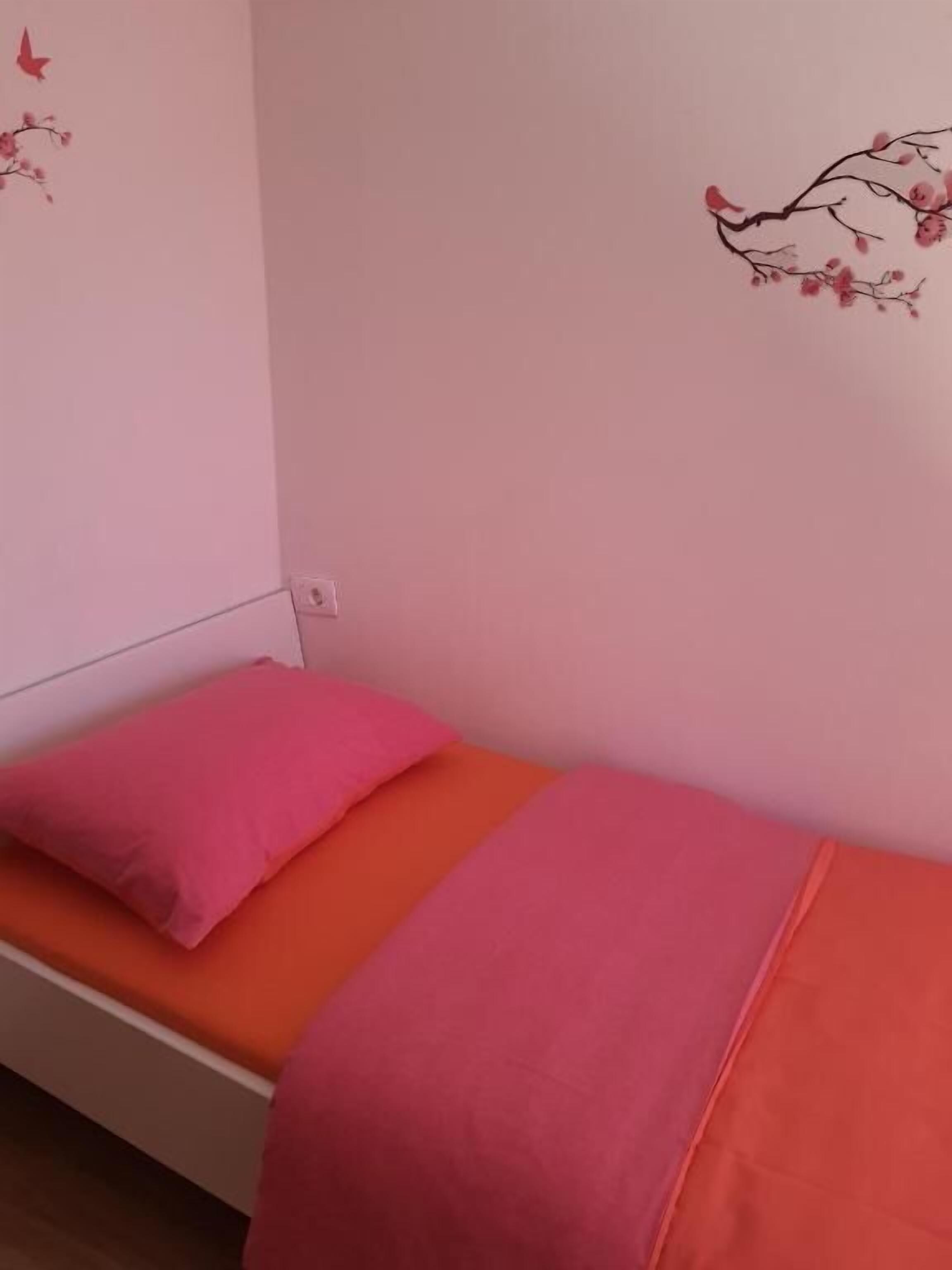 A small, single-bed room with a pink pillow and orange bedding.