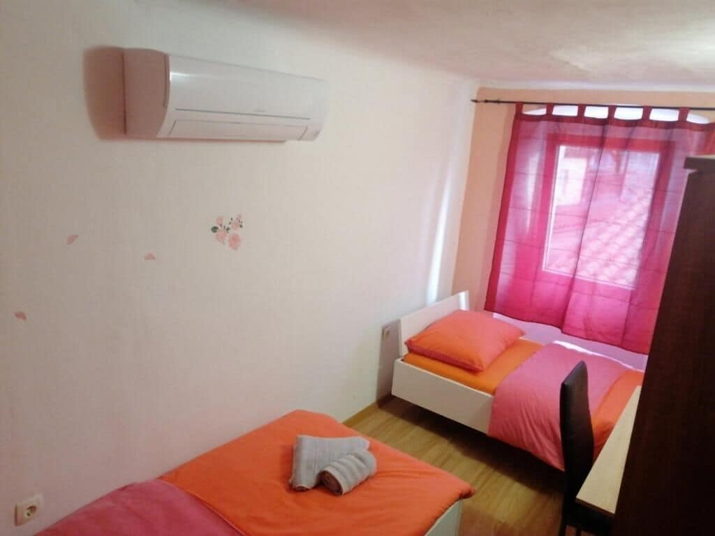 A small, single-room accommodation with two beds, a window with pink curtains, and an air conditioning unit on the wall.