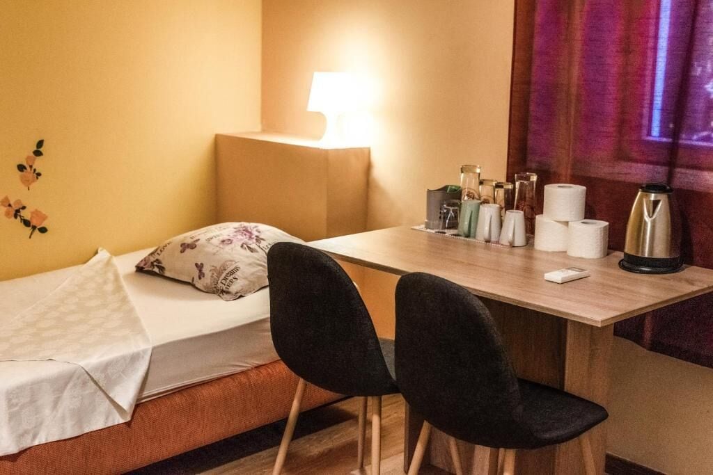 Una habitación de hotel pequeña con una cama individual, un escritorio con lámpara, dos sillas y una ventana con cortinas.