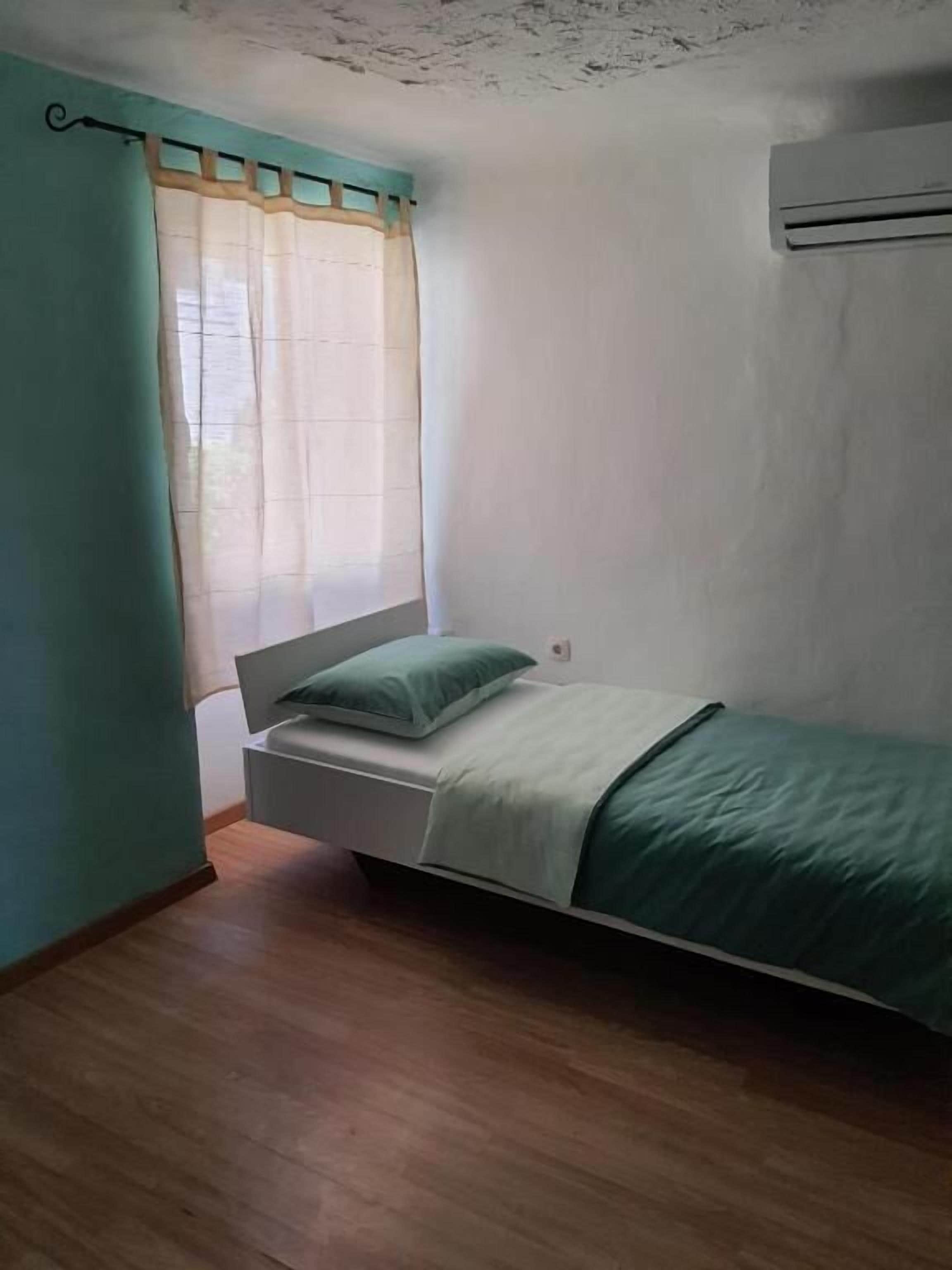 Une petite chambre avec un lit simple, un plancher en bois, une fenêtre avec des rideaux légers et un climatiseur fixé au mur.