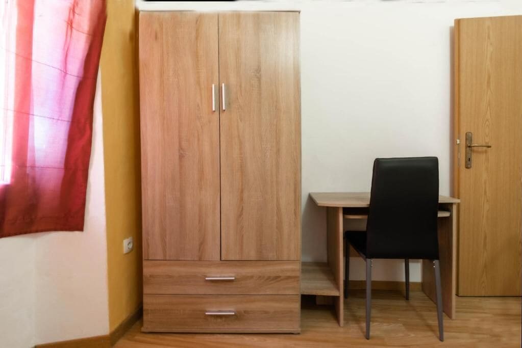 Quarto com guarda-roupa de madeira, escrivaninha com cadeira preta e porta de madeira.